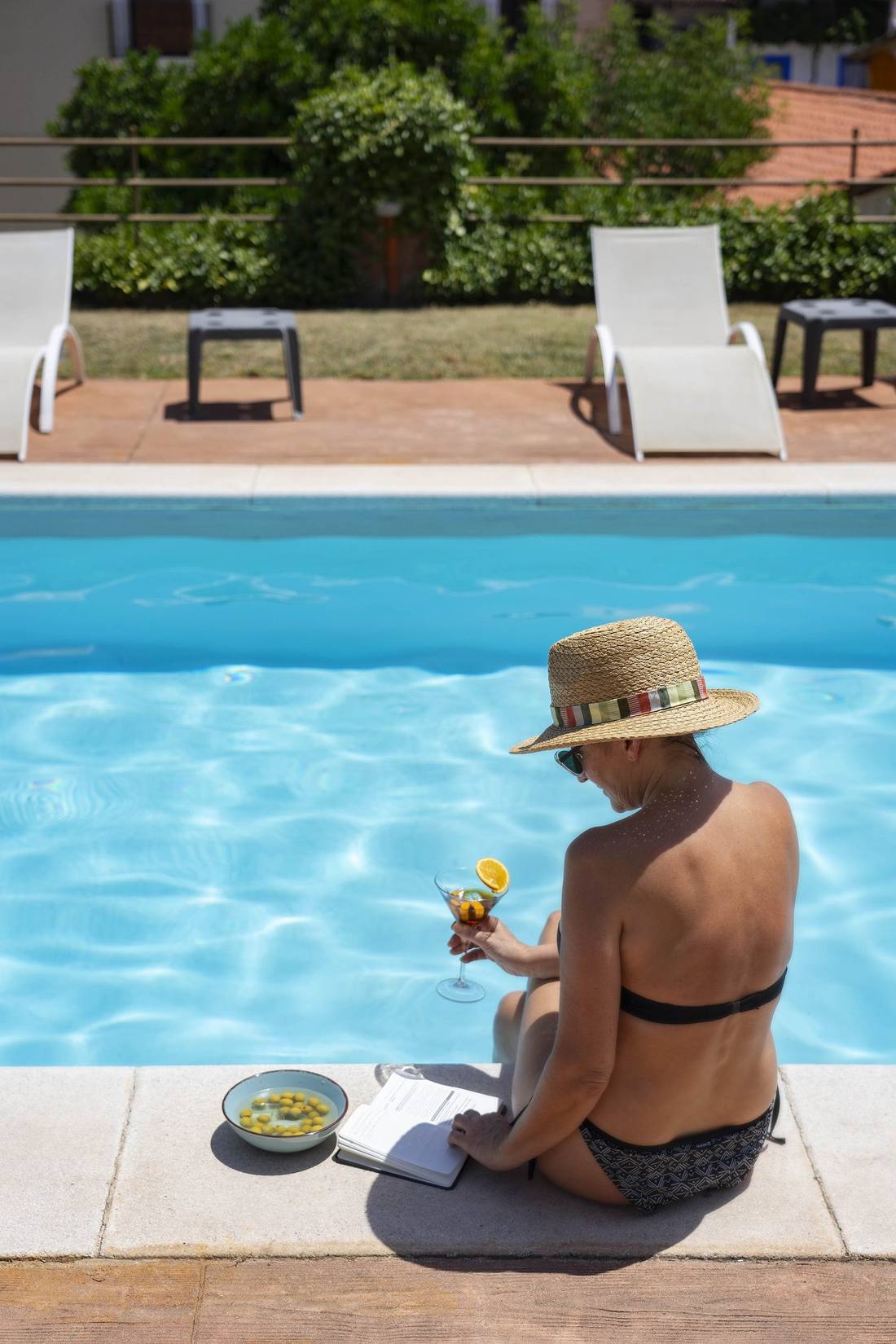 Mujer con sombrero de paja de espaldas en la piscina, sosteniendo una copa